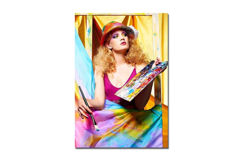 Lerretsbilde 70x100 cm - Kreativ kvinne som maler med en palett i hånden, omgitt av fargerike stoffer, Rosa / Blå / Gul