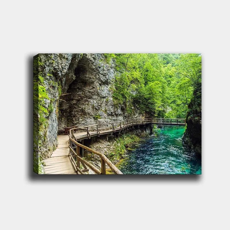 Lerretsbilde 70x100 cm - Naturskjønn utsikt over en trebro som slynger seg gjennom en frodig kløft ved en klar, turkis elv, Grønn / Turkis / Grå