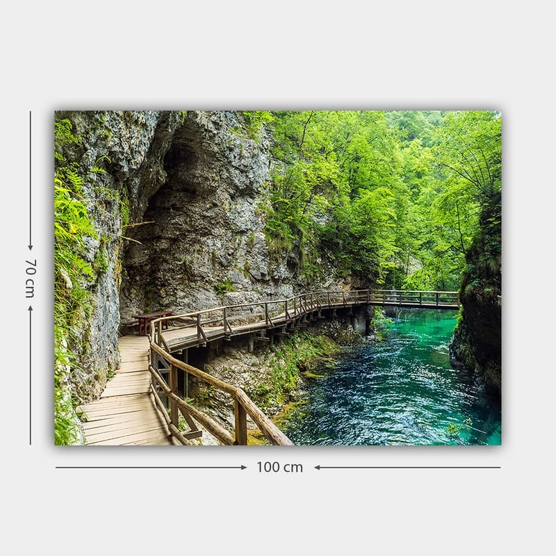 Lerretsbilde 70x100 cm - Naturskjønn utsikt over en trebro som slynger seg gjennom en frodig kløft ved en klar, turkis elv - Grønn / Turkis / Grå - Interiør - Maleri & posters - Lerretsbilder