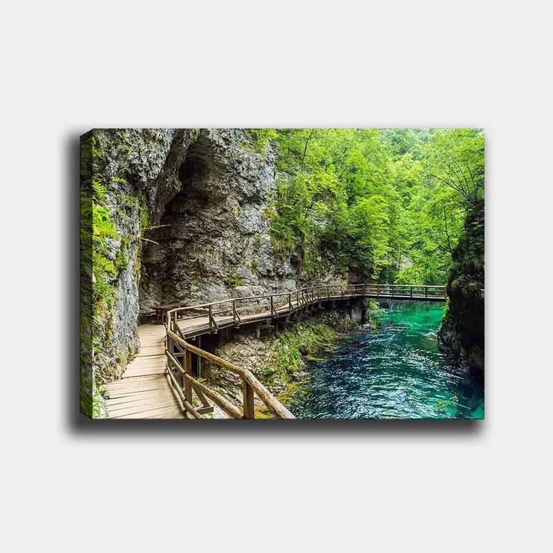 Lerretsbilde 70x100 cm - Naturskjønn utsikt over en trebro som slynger seg gjennom en frodig kløft ved en klar, turkis elv, Grønn / Turkis / Grå