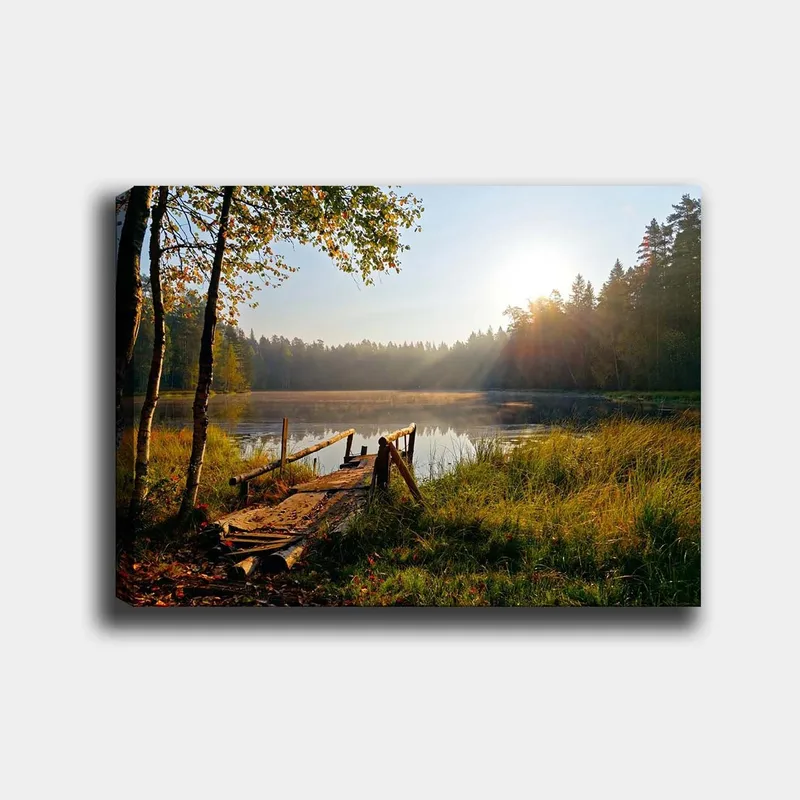 Lerretsbilde 70x100 cm - Rolig innsjø med en trebro omgitt av frodige trær og morgensolens stråler, Grønn / Brun / Gul