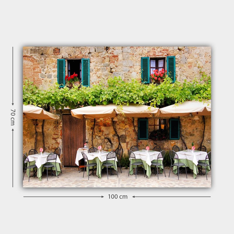 Lerretsbilde 70x100 cm - Sjarmerende utendørs spiseplass med hvite bord og grønne duker foran en gammel steinbygning med mørkegrønne skodder - Mørkegrønn / Hvit / Mursteinsrød - Interiør - Maleri & posters - Lerretsbilder