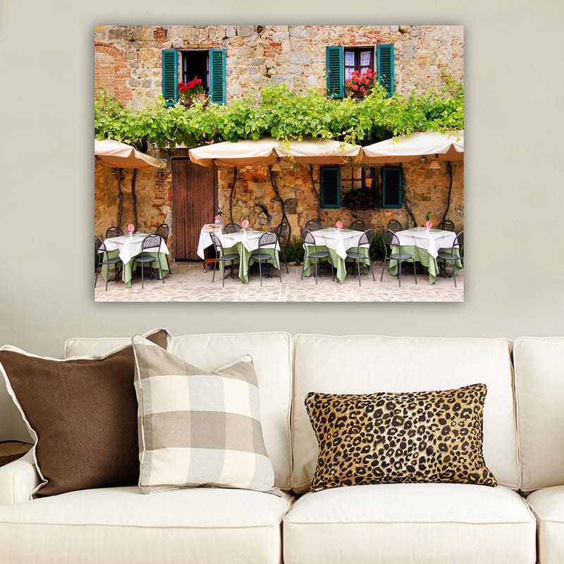Lerretsbilde 70x100 cm - Sjarmerende utendørs spiseplass med hvite bord og grønne duker foran en gammel steinbygning med mørkegrønne skodder - Mørkegrønn / Hvit / Mursteinsrød - Interiør - Maleri & posters - Lerretsbilder