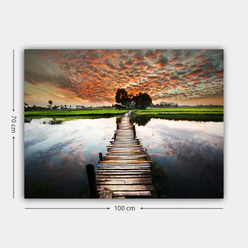 Lerretsbilde 70x100 cm - Trebro som spenner over en speilblank vannoverflate, omgitt av frodig grøntområde og en dramatisk solnedgang - Oransje / Grønn / Blå - Interiør - Maleri & posters - Lerretsbilder