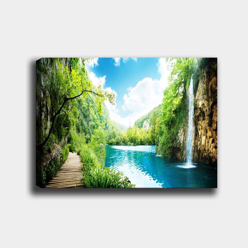 Lerretsbilde 70x100 cm - Vakker naturscene med en foss som renner ut i en klar, blå innsjø omgitt av frodig grøntområde og fjell, Grønn / Blå