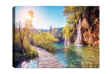 Lerretsbilde 70x100 cm - Vakker naturutsikt med en trebro som fører gjennom frodig grøntområde og en foss i bakgrunnen - Grønn / Blå / Gul - Interiør - Maleri & posters - Lerretsbilder