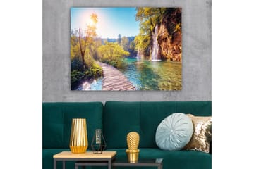 Lerretsbilde 70x100 cm - Vakker naturutsikt med en trebro som fører gjennom frodig grøntområde og en foss i bakgrunnen - Grønn / Blå / Gul - Interiør - Maleri & posters - Lerretsbilder