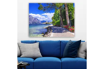 Lerretsbilde 70x100 cm - Vakker promenade med trær og blomster ved en innsjø, omgitt av fjell og en pittoresk by i bakgrunnen - Blå / Grønn / Rød - Interiør - Maleri & posters - Lerretsbilder