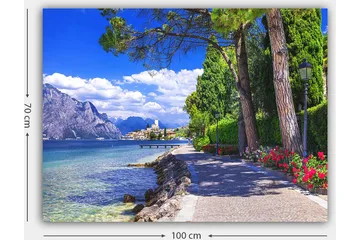 Lerretsbilde 70x100 cm - Vakker promenade med trær og blomster ved en innsjø, omgitt av fjell og en pittoresk by i bakgrunnen - Blå / Grønn / Rød - Interiør - Maleri & posters - Lerretsbilder