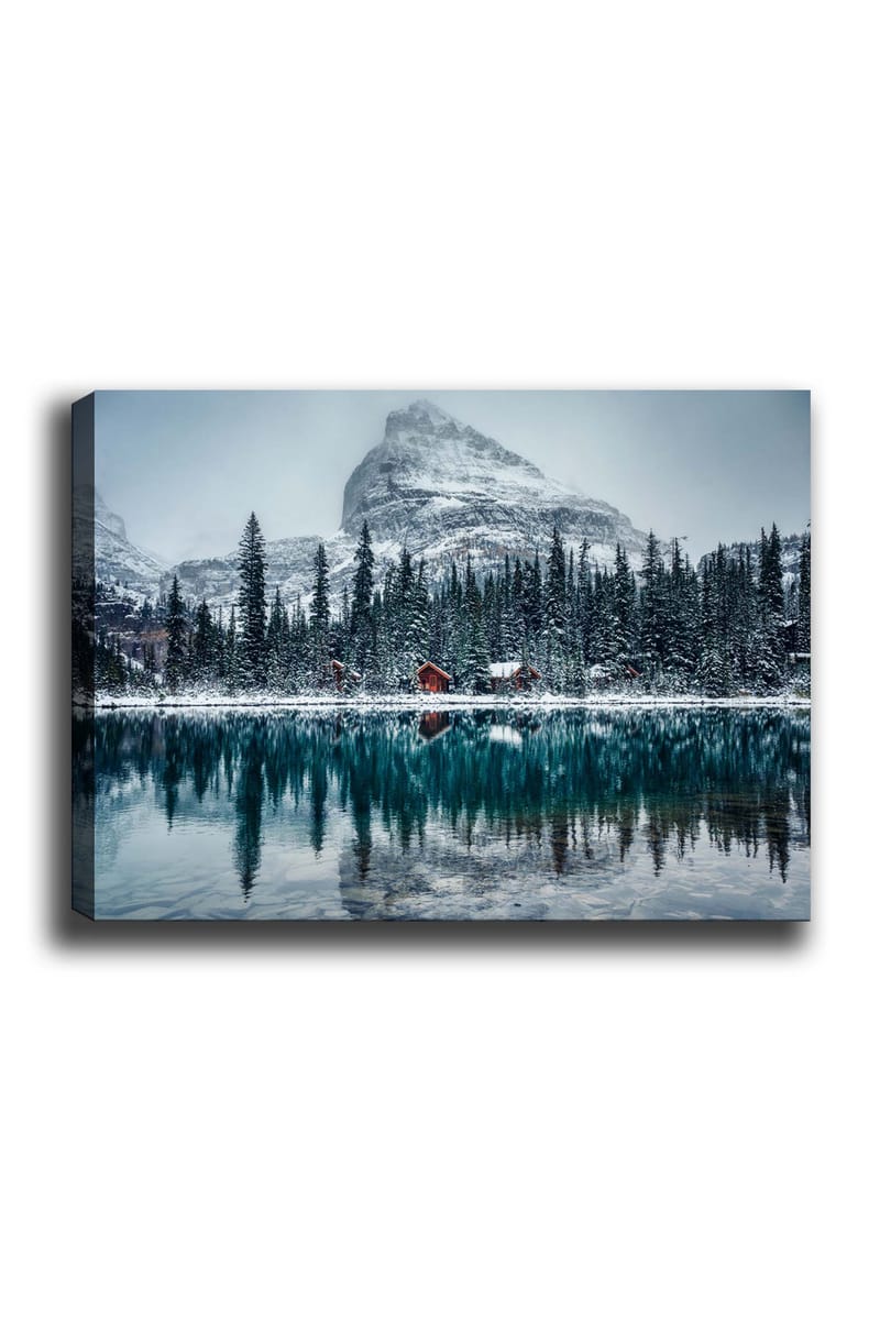 Lerretsbilde 70x100 cm - Vinterlandskap med snødekte fjell og en speilblank innsjø omgitt av grantrær og røde hytter, Mørk grønn / Blå / Hvit