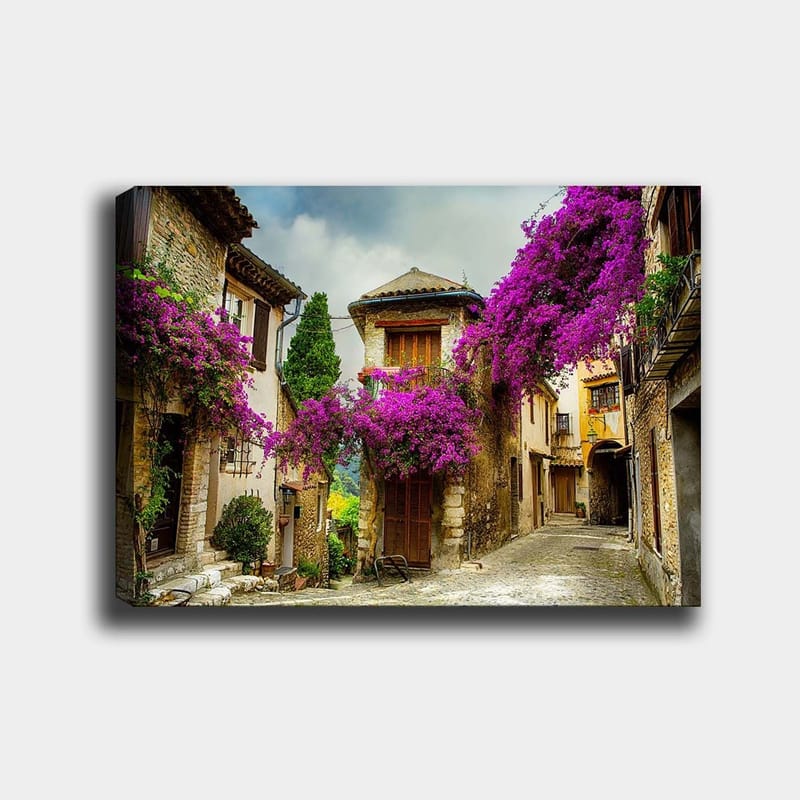 Lerretsbilde 70x100 cm - Pittoresk smug omgitt av fargerike blomster i sjarmerende landsbymiljø, Lilla / Grønn / Beige