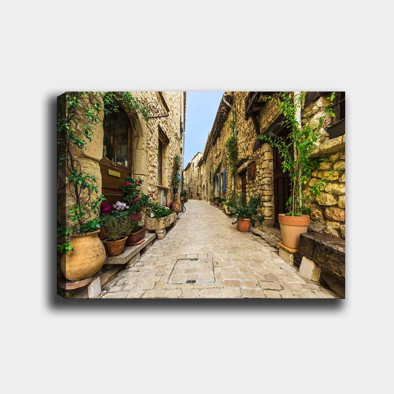 Lerretsbilde 70x100 cm - Pittoresk smug i en gammel landsby med steinhus og blomsterpotter, Beige / Grønn / Terrakotta